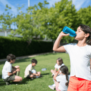 Following cool tips for a healthy summer, a girl drinks from a water bottle in front of friends who are taking a break from playing soccer outside.